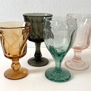 May include: Four stemmed drinking glasses in different colours: amber, green, grey, and pink. The glasses have a textured, faceted design.