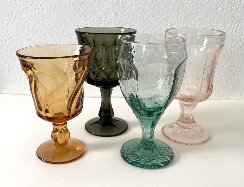 May include: Four stemmed drinking glasses in different colours: amber, green, grey, and pink. The glasses have a textured, faceted design.
