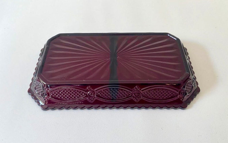 Puede incluir: Un plato de vidrio rojo oscuro con un patr&oacute;n decorativo de rayos de sol en la parte superior y un borde festoneado. El plato tiene una base elevada y con patrones.