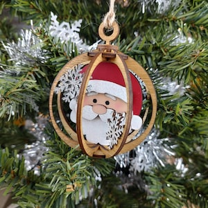 May include: A wooden Christmas ornament shaped like a sphere with a Santa Claus face inside. The ornament is made of multiple layers of wood and has a red hat, white beard, and black eyes. The ornament is hanging on a Christmas tree.
