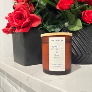 May include: A brown glass candle with a wooden lid. The candle is labeled "DOUCE HAUS" and "ROASTED CHESTNUT & RUM". The candle is handpoured in Brooklyn, NY and made with coconut soy wax.