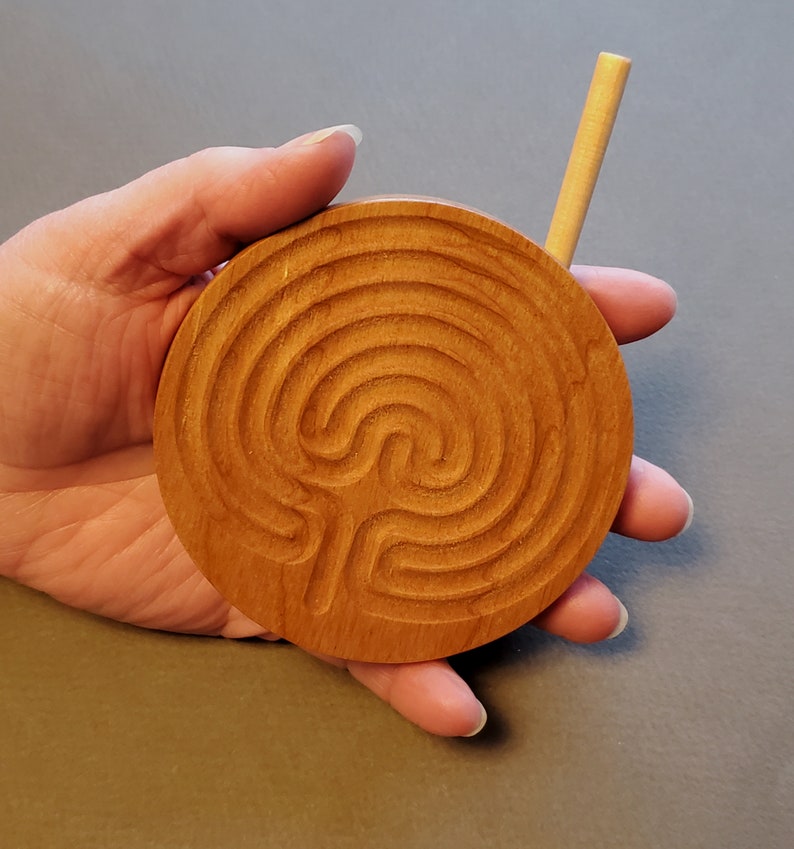 May include: A wooden circular labyrinth with a carved design. A small wooden stick is used to trace the path of the labyrinth.