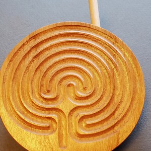 May include: A wooden circular labyrinth with a carved design. A wooden stick is placed on top of the labyrinth.