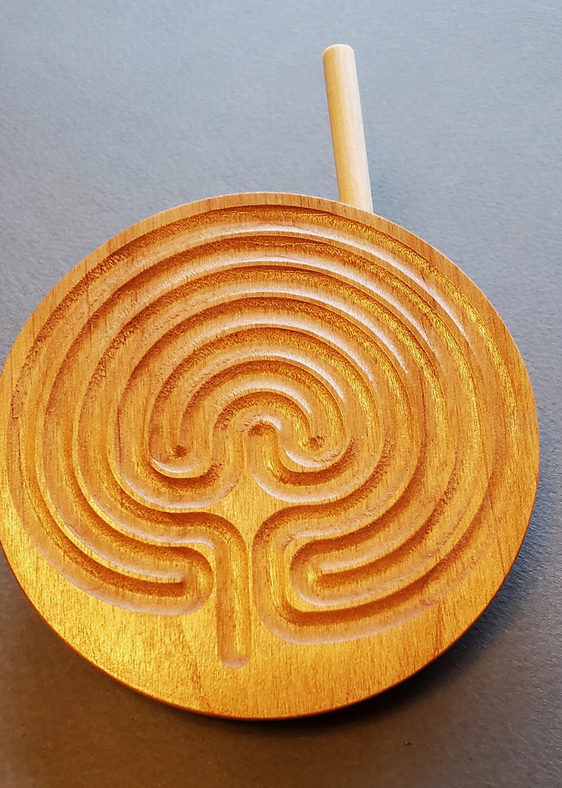 May include: A wooden circular labyrinth with a carved design. A wooden stick is placed on top of the labyrinth.