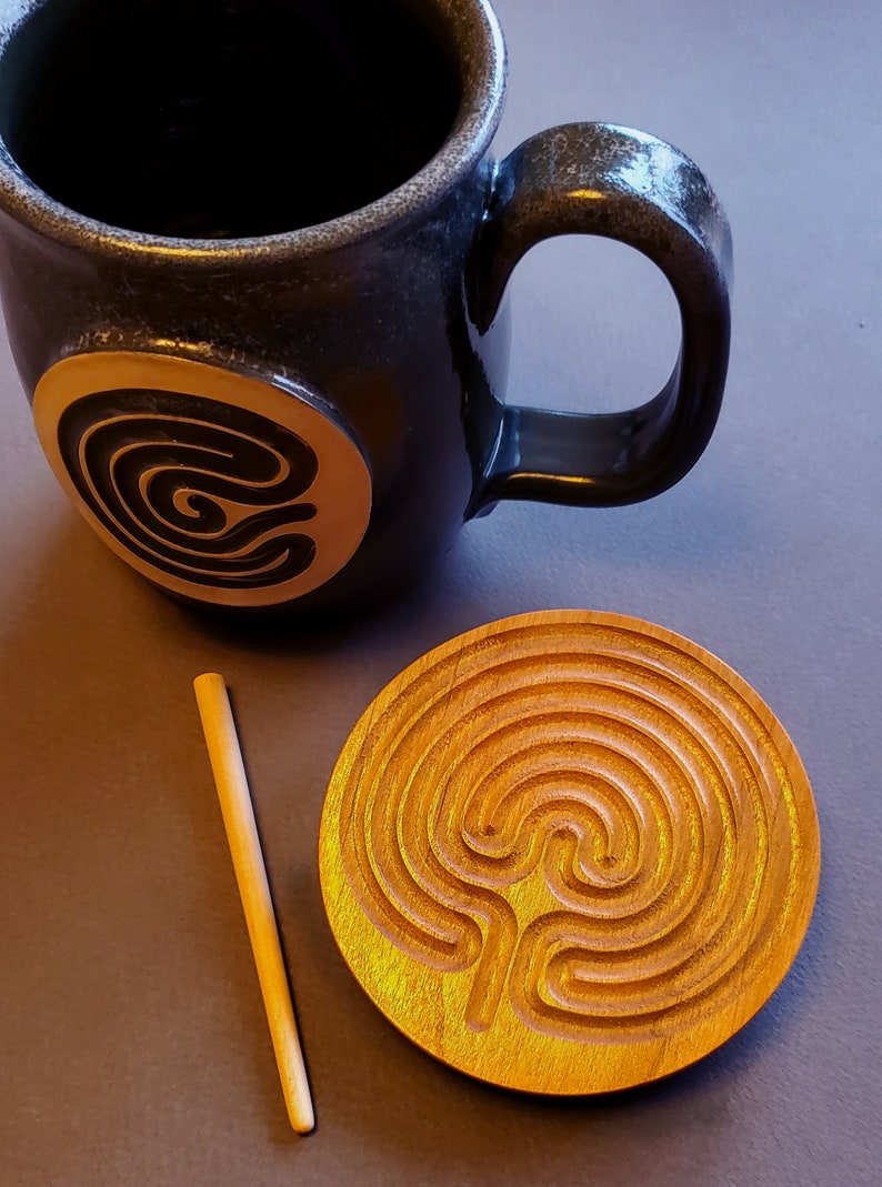 May include: A wooden labyrinth coaster with a light brown finish. The coaster features a carved labyrinth design with a central circle. A wooden stick is also included.