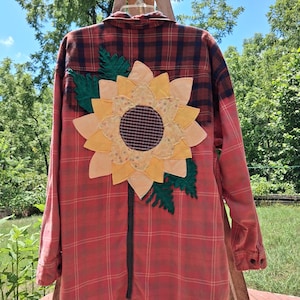 May include: A long-sleeve button-down shirt in a red and black plaid pattern featuring a large fabric sunflower appliqué on the back. The sunflower has yellow petals, a dark brown center, and dark green fabric leaves attached to the shirt.