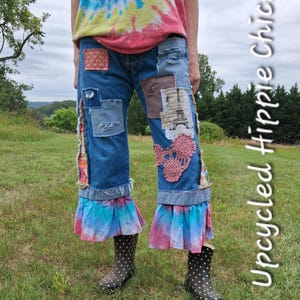 May include: Blue denim jeans featuring various fabric patches, including a square with an Eiffel Tower print, a pink crocheted floral applique, and frayed edges. The hem is finished with a ruffled, tie-dye fabric in pink and blue tones.