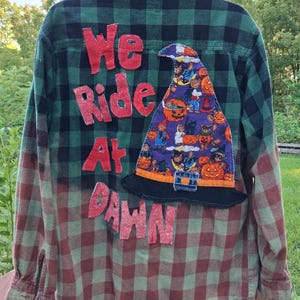 May include: A long-sleeve button-down flannel shirt featuring a green and black plaid pattern with a faded red lower section. The back displays red fabric letters spelling "We Ride At Dawn" and a witch hat patch with a purple Halloween print.