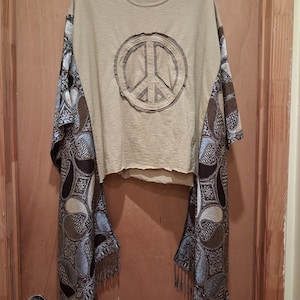 May include: A tan short-sleeved t-shirt with a raw-edge hem and a central peace sign graphic. The sides feature long, draped panels of dark brown and light blue fabric with a detailed paisley pattern and fringed edges at the bottom hem.
