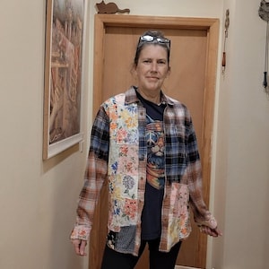 May include: A long-sleeved button-down shirt featuring a patchwork design of plaid and floral fabric panels in shades of blue, brown, and pink, worn over a dark t-shirt with a cat graphic, paired with black pants and a sign reading Upcycled Hippie Chick.