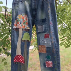 May include: Blue denim jeans featuring fabric mushroom appliques on the right leg and various colorful patches on the left leg. The text "TAKE A LITTLE TRIP" is embroidered vertically down the left side in multi-colored thread.