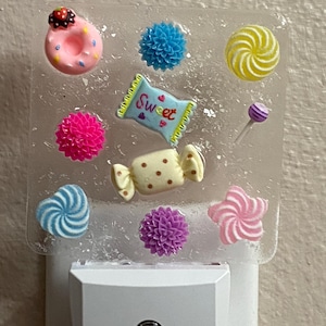May include: A decorative night light featuring various candy-themed embellishments. The clear plastic base is adorned with a pink donut, blue and pink pom-poms, a candy wrapper with the word "Sweet", and other colorful sweets.