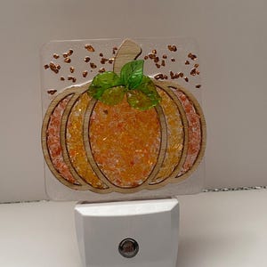 May include: A night light featuring a square clear acrylic panel with a pumpkin design. The pumpkin is outlined in wood with orange and yellow crushed glass fill, topped with two green leaves. Copper-colored flakes are embedded in the clear panel.