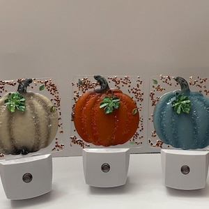 May include: Three glass night lights featuring pumpkin designs in beige, orange, and blue. Each pumpkin has a green leaf, a dark stem, and is set against a square clear glass background decorated with small copper-colored metallic flakes.