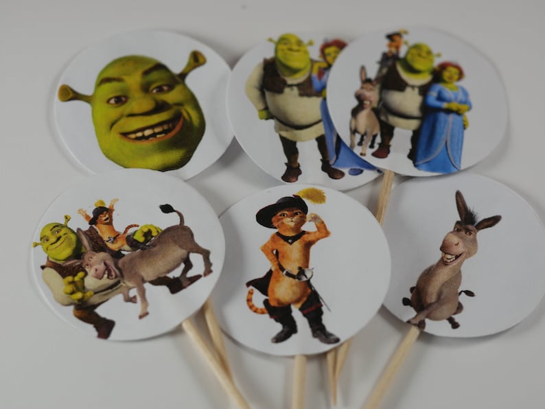May include: Set of 6 round cupcake toppers featuring characters from the Shrek movie franchise. The toppers feature Shrek, Fiona, Donkey, Puss in Boots, and a close-up of Shrek's face.
