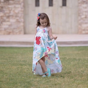 Peut inclure: Une jeune fille porte une robe bleu clair asymétrique avec un imprimé coloré d'Ariel la Petite Sirène. Elle porte également des chaussures Mary Jane bleu clair et un nœud coloré dans les cheveux. La fille est sur l'herbe verte.