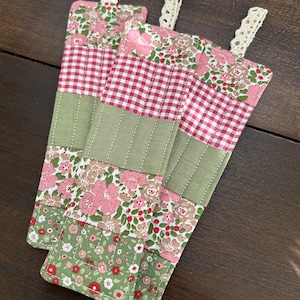 Op de afbeelding: Drie handgemaakte stoffen bladwijzers met een patchwork-ontwerp. De bladwijzers hebben een groen, roze en rood bloemenpatroon met witte gingham.
