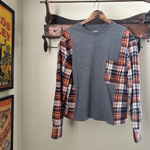 Könnte beinhalten: Ein graues Langarmshirt mit karierten Ärmeln und einer Tasche. Das Karomuster umfasst Farbtöne von Orange, Braun, Weiß und Blau. Das Shirt hängt an einem Holzbügel.