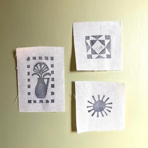 May include: Three rectangular and square pieces of off-white fabric featuring dark gray block-printed designs. One piece shows a vase with a plant, another a geometric square pattern, and the third a circular sun-like motif with radiating rays.