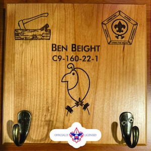 Puede incluir: Una placa de madera con dos ganchos, un emblema de scout y el texto "BEN BEIGHT C9-160-22-1 OFFICIALLY LICENSED".