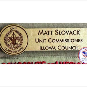 May include: A light-colored wooden name tag featuring a circular emblem with the text UNIT COMMISSIONER and a central fleur-de-lis. The rectangular portion displays the engraved text MATT SLOVACK, UNIT COMMISSIONER, and ILLOWA COUNCIL.