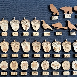 May include: A collection of 37 wooden acorn and round shaped engraved badges. The badges feature various designs including animals, scout emblems, and text. The badges are arranged in rows on a dark blue surface.