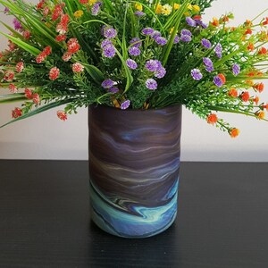 May include: A decorative vase with a swirling, abstract design in shades of blue, brown, and purple. The vase is filled with a vibrant arrangement of artificial flowers in various colors, including red, purple, yellow, and orange, with green grass.