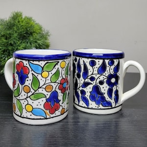 May include: Two white ceramic mugs with blue, green, red and yellow floral designs. The mugs have a blue rim and a handle.