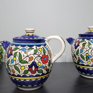 Puede incluir: Dos teteras de cerámica con tapas, con una base blanca y intrincados diseños florales en rojo, azul, verde y amarillo. Las teteras tienen bordes y asas azules, y se exhiben sobre una superficie oscura.