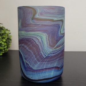 May include: A cylindrical glass vase with a marbled design. The vase features a blend of blue, purple, and green hues, creating a layered, organic pattern. The vase is set against a neutral background.