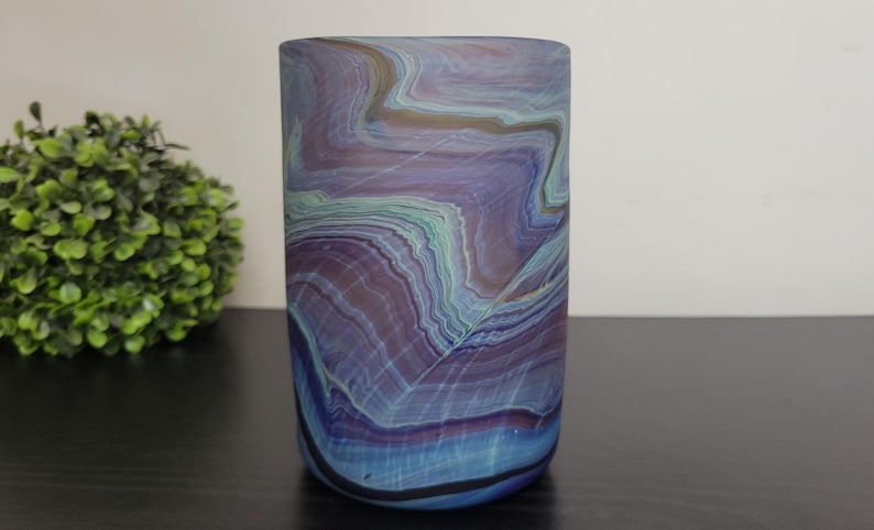 May include: A cylindrical glass vase with a marbled design. The vase features a blend of blue, purple, and green hues, creating a layered, organic pattern. The vase is set against a neutral background.