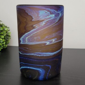 May include: A cylindrical glass vase with a swirling pattern of brown, blue, and purple. The colors blend together, creating a marbled effect. The vase is on a dark surface.