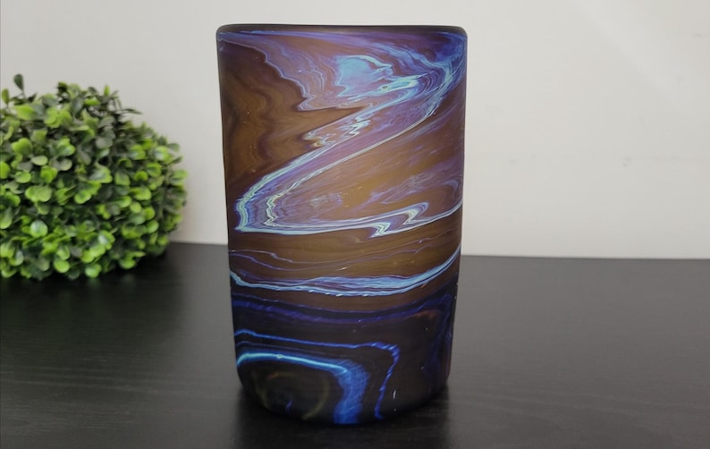 May include: A cylindrical glass vase with a swirling pattern of brown, blue, and purple. The colors blend together, creating a marbled effect. The vase is on a dark surface.