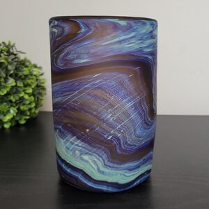 May include: A cylindrical glass vase with a swirling pattern of blue, purple, and brown colors. The vase has a smooth surface and is set against a neutral background. The design resembles agate stone.