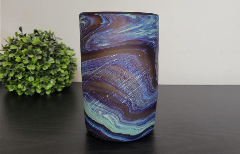 May include: A cylindrical glass vase with a swirling pattern of blue, purple, and brown colors. The vase has a smooth surface and is set against a neutral background. The design resembles agate stone.