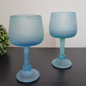 Könnte beinhalten: Zwei blau getönte Weingläser mit mattierter Oberfläche. Die Gläser haben eine abgerundete Schalenform und stehen auf einem Stiel mit dekorativem Fuß. Das Glas hat ein marmoriertes Aussehen mit helleren und dunkleren Blautönen.