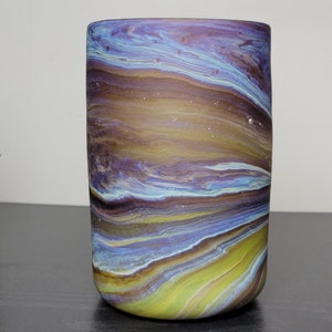 May include: A cylindrical glass vase with a swirling pattern of colors. The design features shades of blue, yellow, and brown, creating a marbled effect. The vase is approximately 6 inches tall and has a smooth, rounded top.