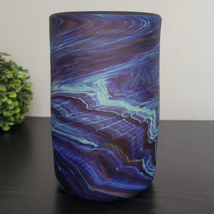 May include: A cylindrical glass vase with a swirling pattern of deep blue, purple, and teal colors. The vase has a smooth, polished finish and stands upright on a dark surface. A green, spherical plant is in the background.