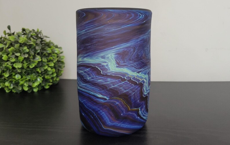 May include: A cylindrical glass vase with a swirling pattern of deep blue, purple, and teal colors. The vase has a smooth, polished finish and stands upright on a dark surface. A green, spherical plant is in the background.