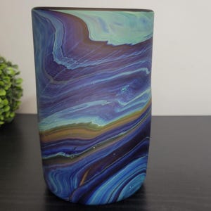 May include: A cylindrical glass vase with a swirling pattern of blue, purple, and teal colors. The vase has a smooth, glossy finish and is displayed on a dark surface. The design resembles a watercolor painting.