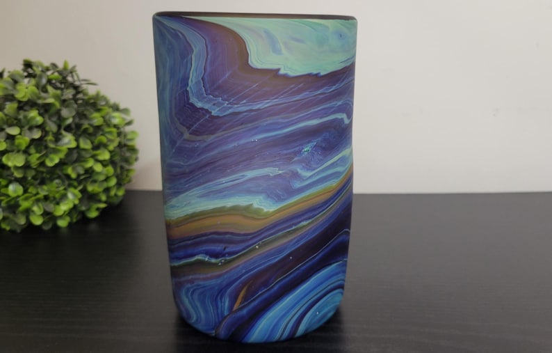 May include: A cylindrical glass vase with a swirling pattern of blue, purple, and teal colors. The vase has a smooth, glossy finish and is displayed on a dark surface. The design resembles a watercolor painting.