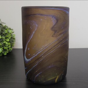May include: A cylindrical vase with a swirling pattern of brown, blue, and gold. The vase is made of glass and has a smooth, polished surface. The vase is approximately 6 inches tall.