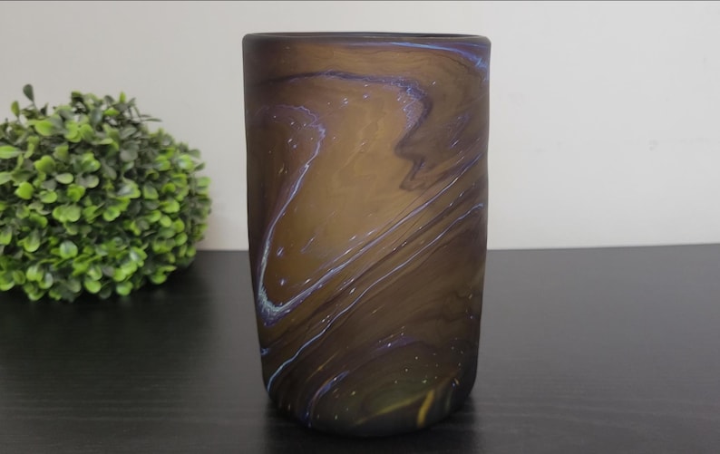 May include: A cylindrical vase with a swirling pattern of brown, blue, and gold. The vase is made of glass and has a smooth, polished surface. The vase is approximately 6 inches tall.