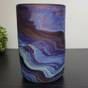 May include: A cylindrical glass vase with a swirling pattern of blue, purple, and brown hues. The vase has a frosted finish and sits on a dark surface. A green, spherical plant is in the background.