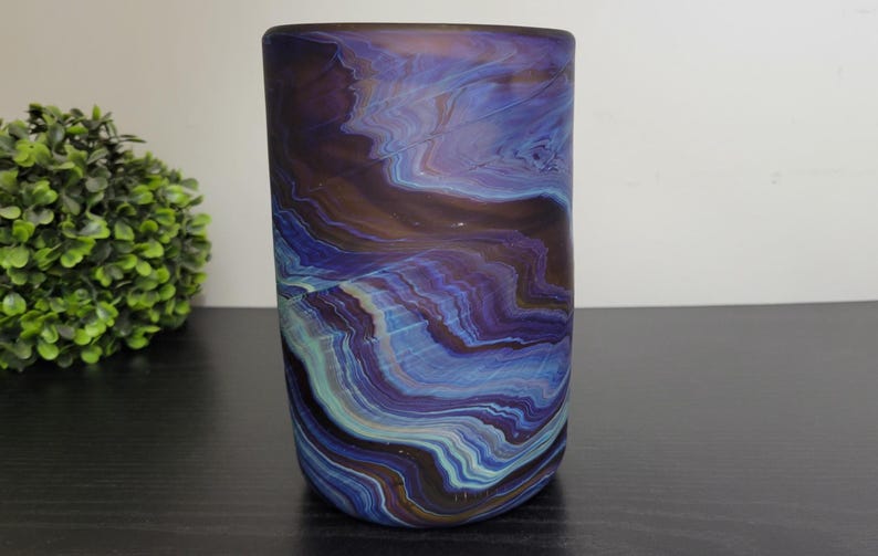 May include: A cylindrical glass vase with a swirling pattern of blue, purple, and brown hues. The vase has a frosted finish and sits on a dark surface. A green, spherical plant is in the background.