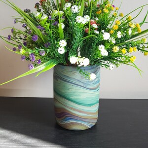 May include: A decorative floral arrangement in a cylindrical vase. The vase has a marbled pattern in shades of blue, green, and brown. The arrangement features a variety of colorful artificial flowers and greenery, including white, yellow, red, and purple.