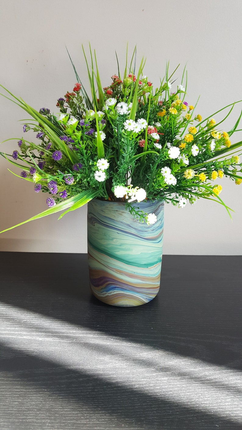 May include: A decorative floral arrangement in a cylindrical vase. The vase has a marbled pattern in shades of blue, green, and brown. The arrangement features a variety of colorful artificial flowers and greenery, including white, yellow, red, and purple.
