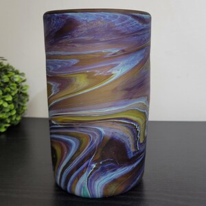 May include: A cylindrical art glass vase with a swirling pattern of blue, purple, yellow, and brown. The vase has a smooth, polished finish and a dark brown rim. The vase is a decorative home accent.