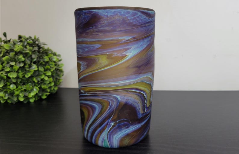 May include: A cylindrical art glass vase with a swirling pattern of blue, purple, yellow, and brown. The vase has a smooth, polished finish and a dark brown rim. The vase is a decorative home accent.