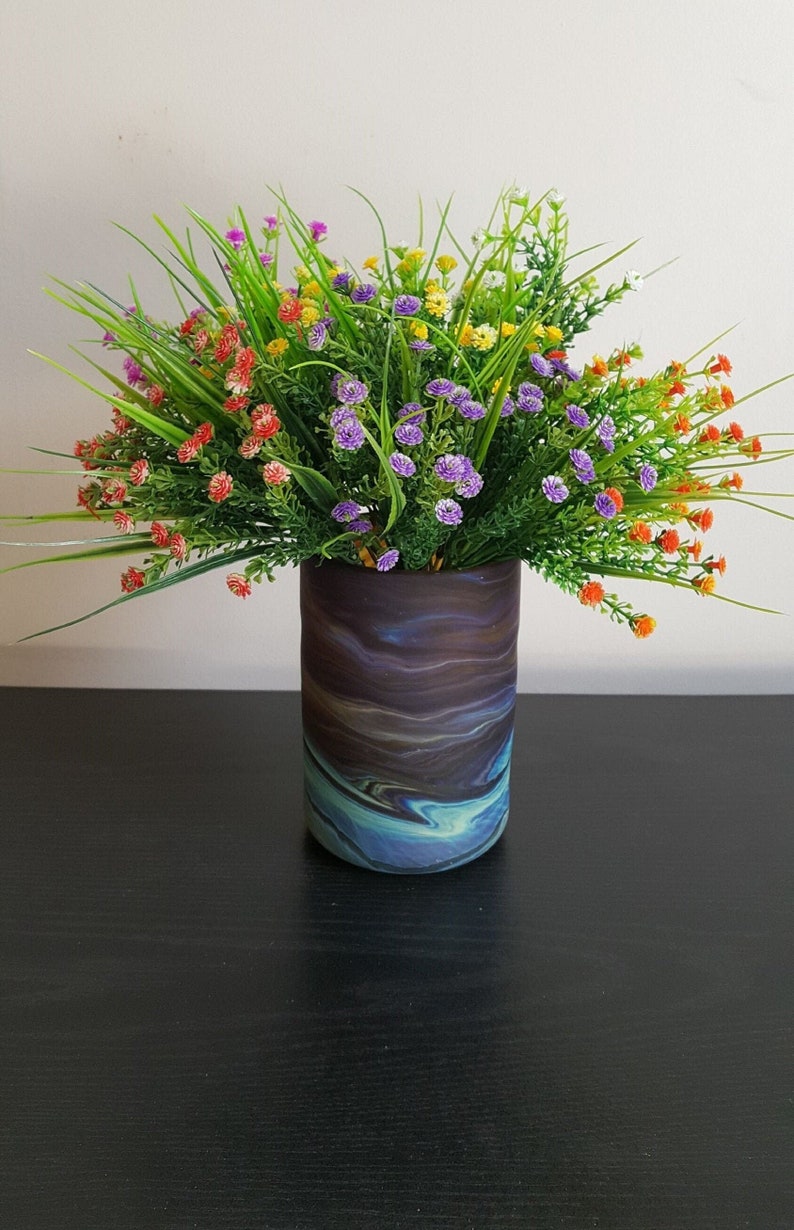 May include: A decorative vase with a swirling, abstract design in shades of blue, brown, and purple. The vase is filled with a vibrant arrangement of artificial flowers in various colors, including red, purple, yellow, and orange, with green grass.